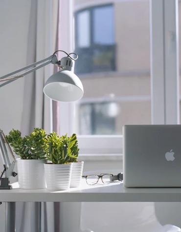 desk with computer and lamp on it with a window blurred out behind it. potted plants sit on the table beside a pair of glasses