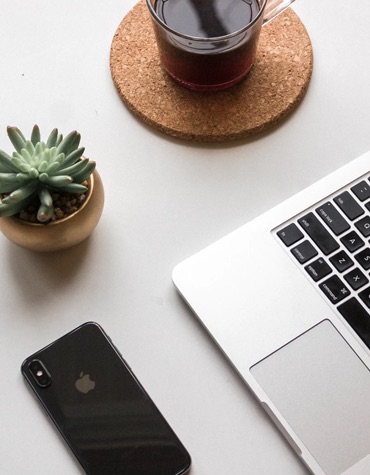 tabletop with a phone upside down a plant, a beverage on a coaster, and an open laptop